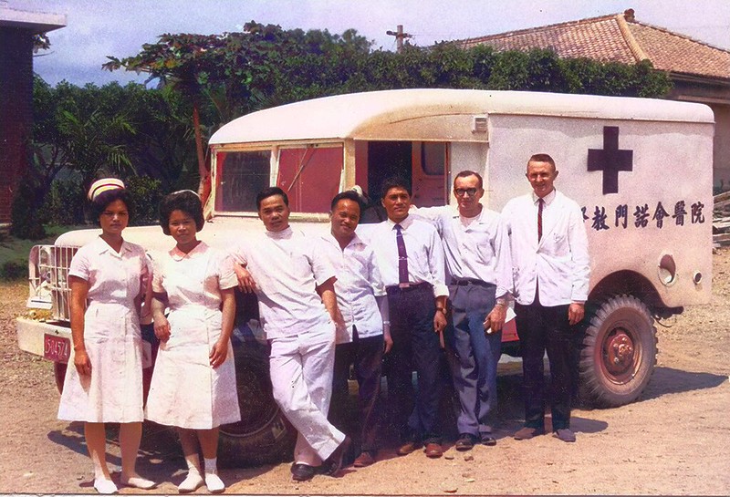 一群醫療工作者站在一輛標有「門諾醫院」的老式救護車旁，背景為台灣的鄉村風景，展現他們的醫療奉獻精神。