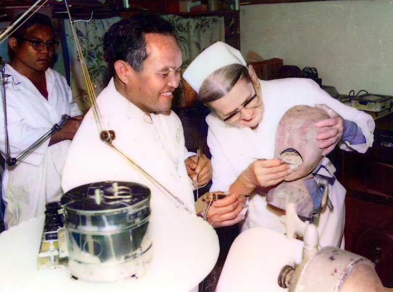 一位醫生與一位女性修女正在討論和調整一個醫療器械，背景中有其他醫療工作人員和設備。