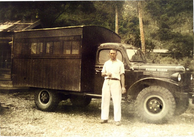 一位男性站在一輛老式卡車旁，背景為樹木和簡易建築，展現早期醫療工作的場景。