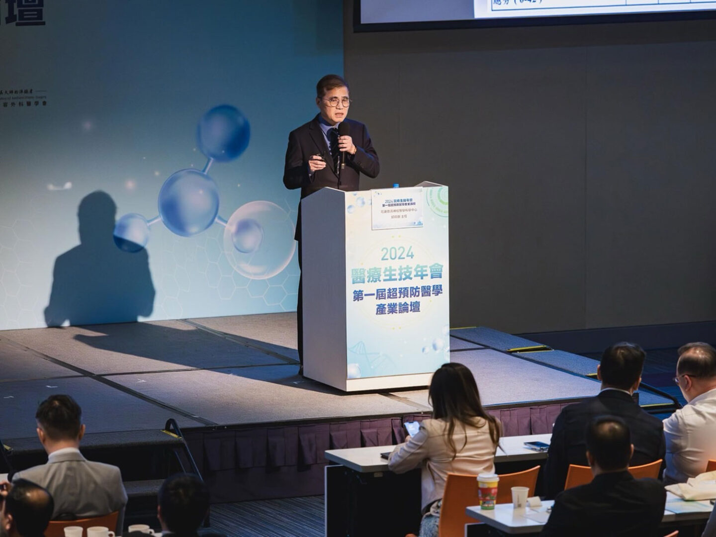 邱琮朗醫師在2024醫療生技年會上發表演講,背景有科學圖形,與會者專注聆聽。
