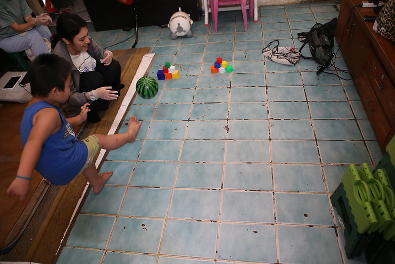 A child in a blue shirt plays on a tiled floor, engaging with a therapist who is encouraging him, while colorful building blocks are arranged nearby.