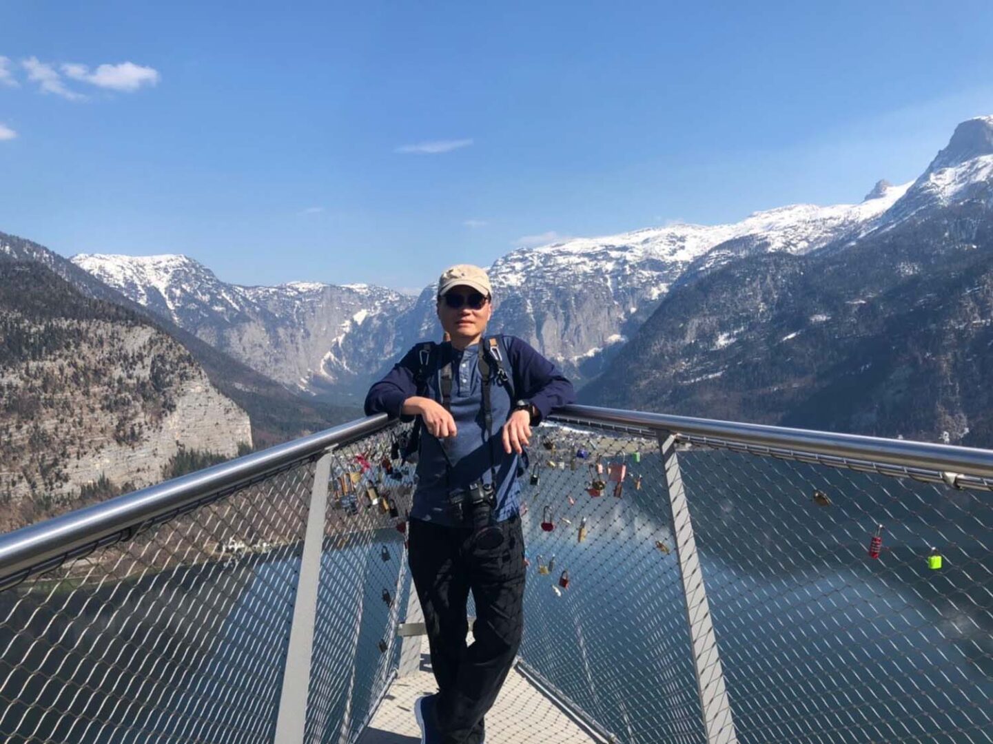 一位男子站在山谷中的金屬懸索橋上,俯瞰峽谷和湖泊,背景是雪山和清晰的藍天。