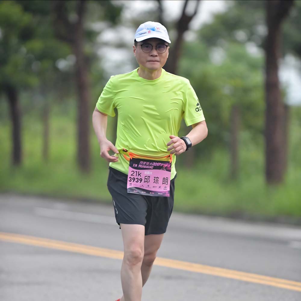 一位穿著明亮綠色運動衫和黑色短褲的男性在馬路上跑步,展示他參加21公里賽跑的紀念牌,背景為綠色植物和樹木。